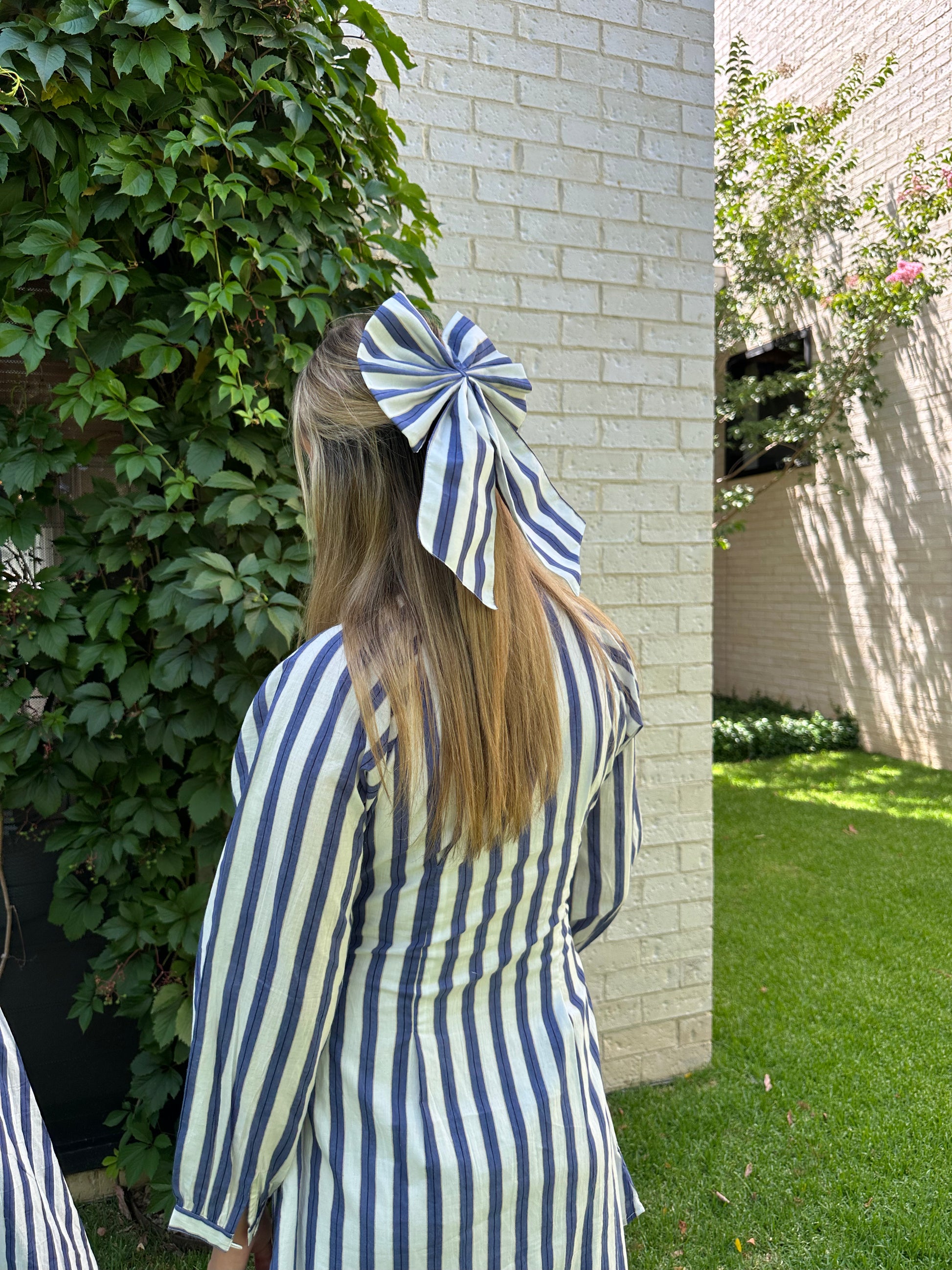 Person wearing a blue and white striped outfit with a large bow in their hair, standing against a brick wall.