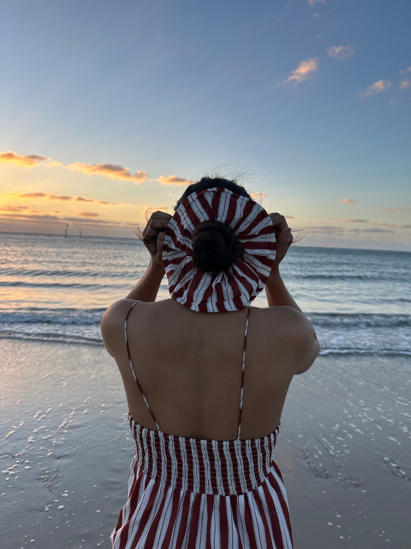 Person in a striped outfit holding a red and white patterned hat on a beach at sunset.