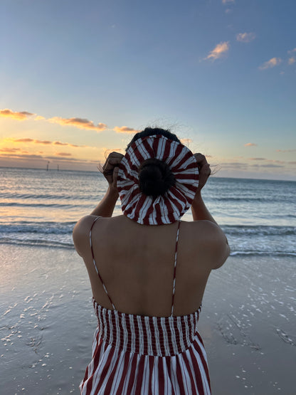 Person in a striped outfit holding a red and white patterned hat on a beach at sunset.
