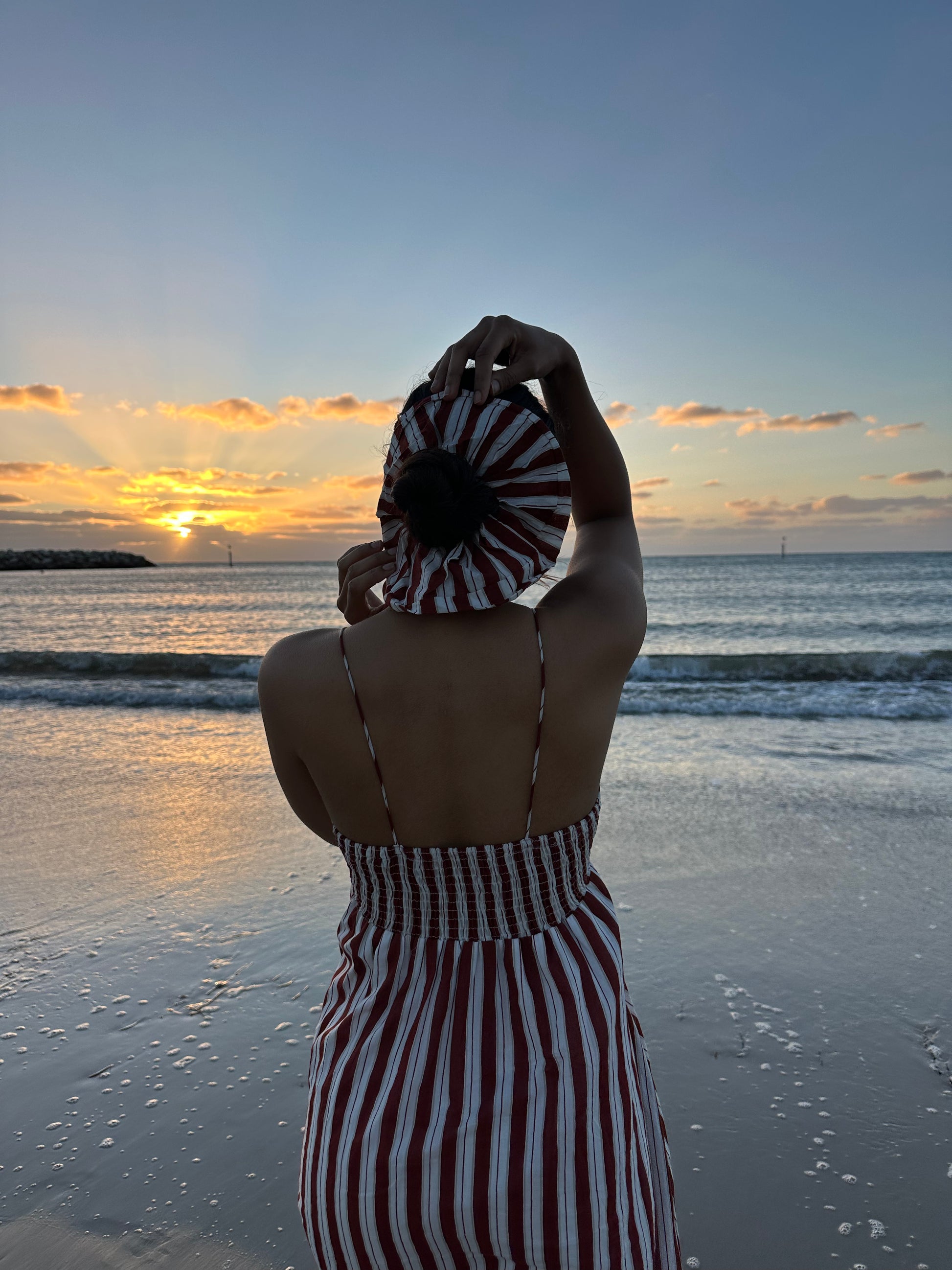 Person in a striped dress holding a hood over their head on a beach at sunset.