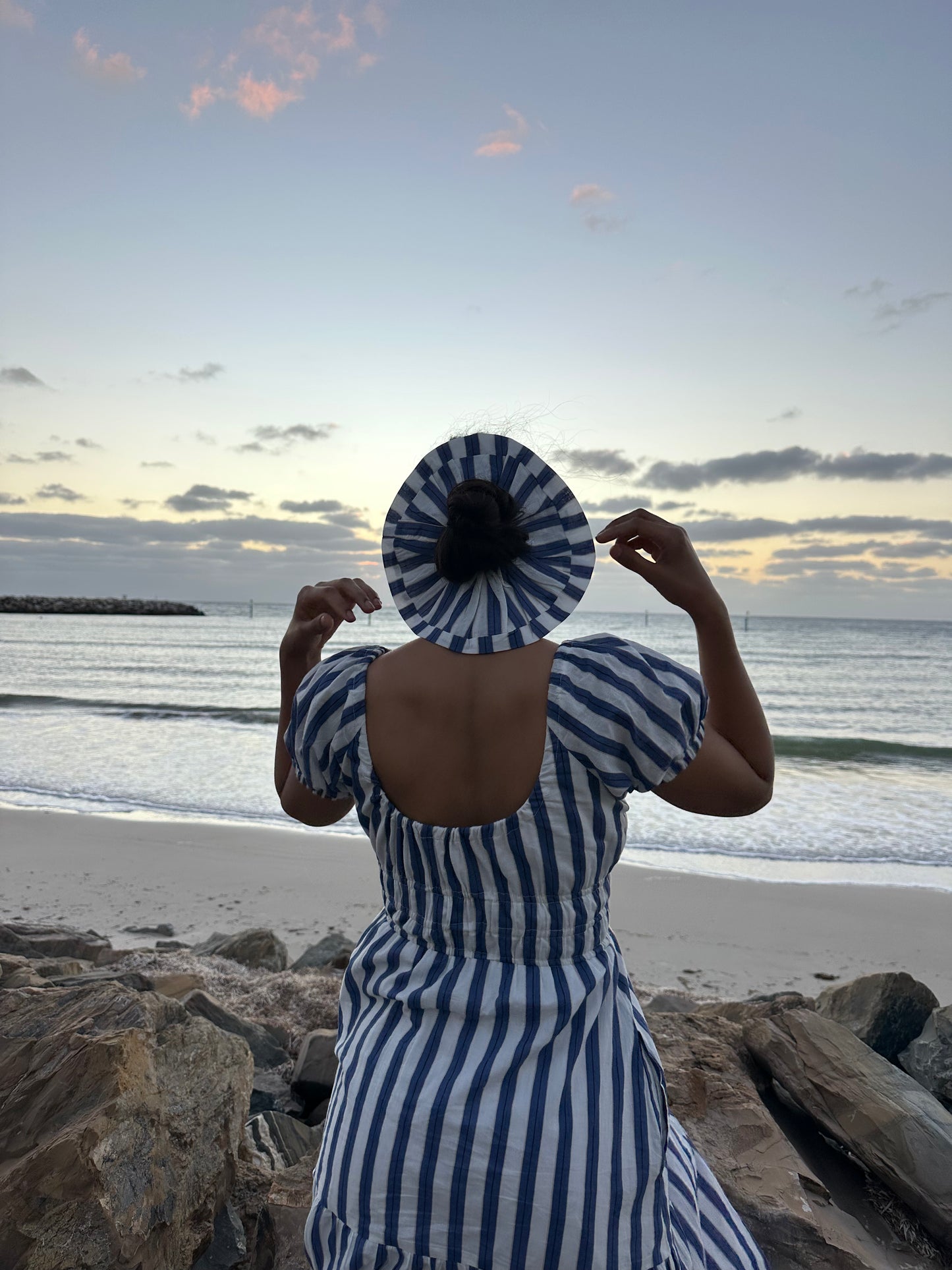Person in a blue and white striped dress and hat standing on a beach at sunset.