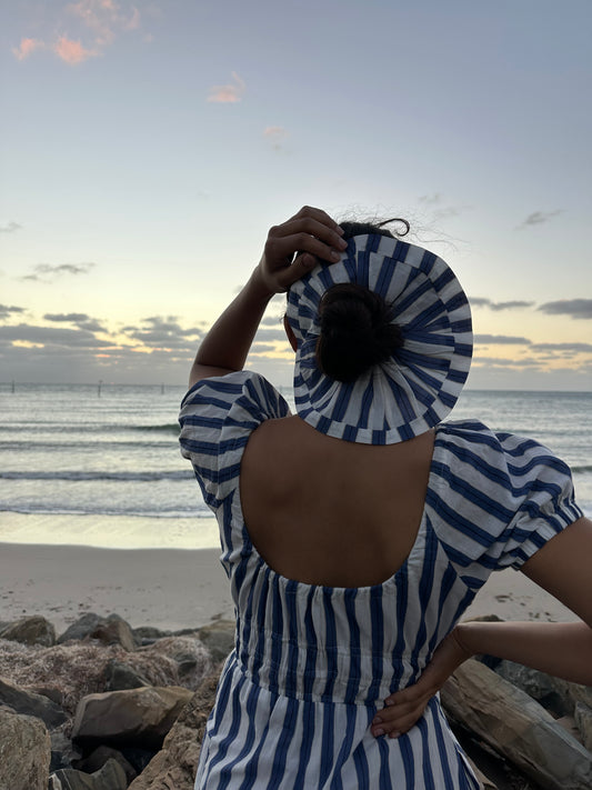 Person wearing a blue and white striped dress and hat on a beach at sunset.