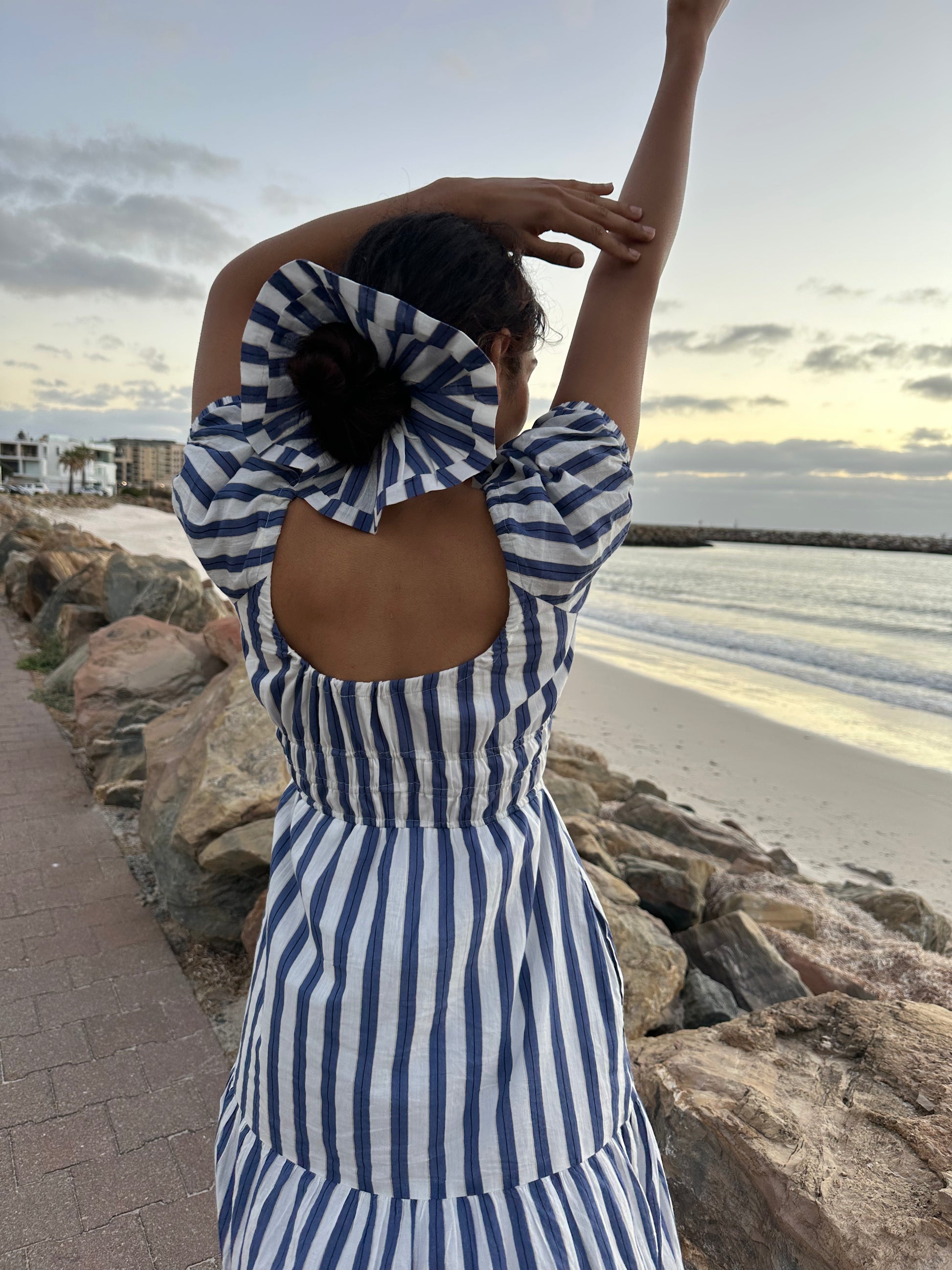 Woman in a blue and white striped dress standing on a beach with a sunset in the background.